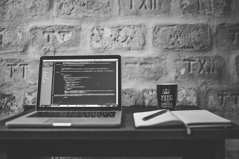 A desktop setup on a dark wooden desk featuring an open laptop displaying lines of code on a black background, with a white notebook and a black pen placed beside it. To the right of the laptop, there